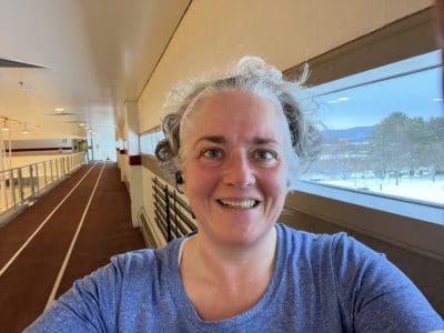 Gray haired woman in a blue shirt on an indoor track. A snowy landscape is in the window over her left shoulder.