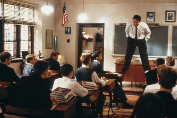 A man standing on a desk addressing a classroom of students