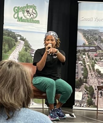 A Black woman speaking into a microphone in front of a Grandma's Marathon banner.