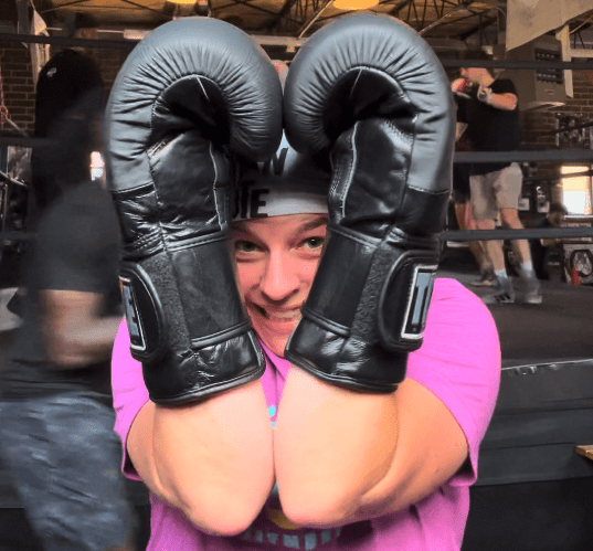 Woman holding boxing gloves up around her face to form a heart with her smiling face in the center.